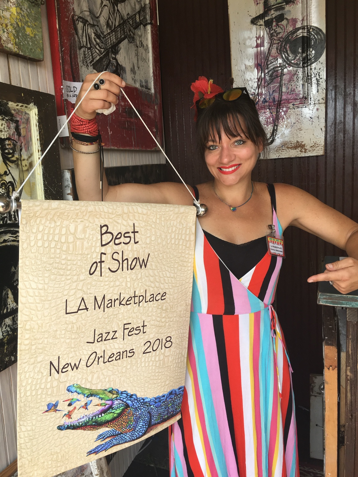 Crystal Obeidzinski smiling and pointing to a cream banner she holds that reads Best of Show, LA Marketplace, Jazz Fest, New Orleans 2018, with her paintings on the wall behind her.