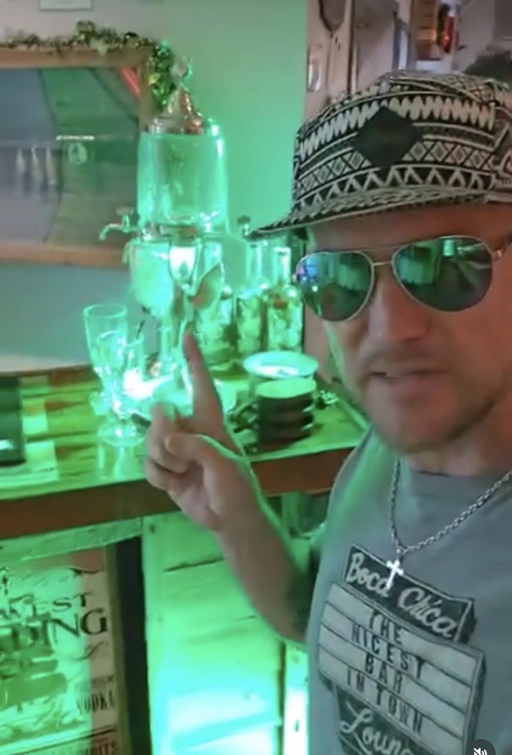 A man in a patterned hat and sunglasses pointing at glassware lit in green.