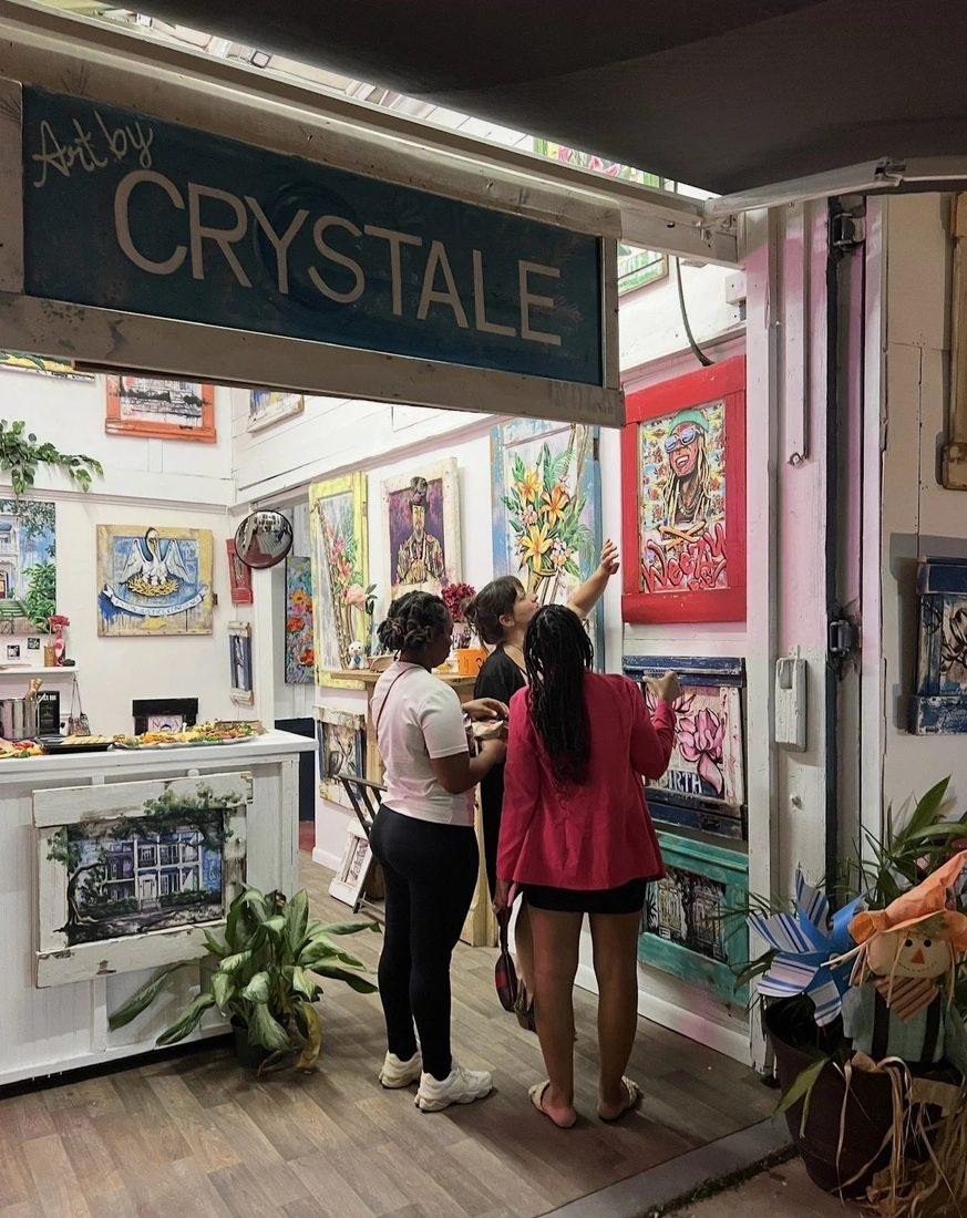 The side entrance to Art by Crystale, a sign over a doorway into a room hung with colorful paintings.