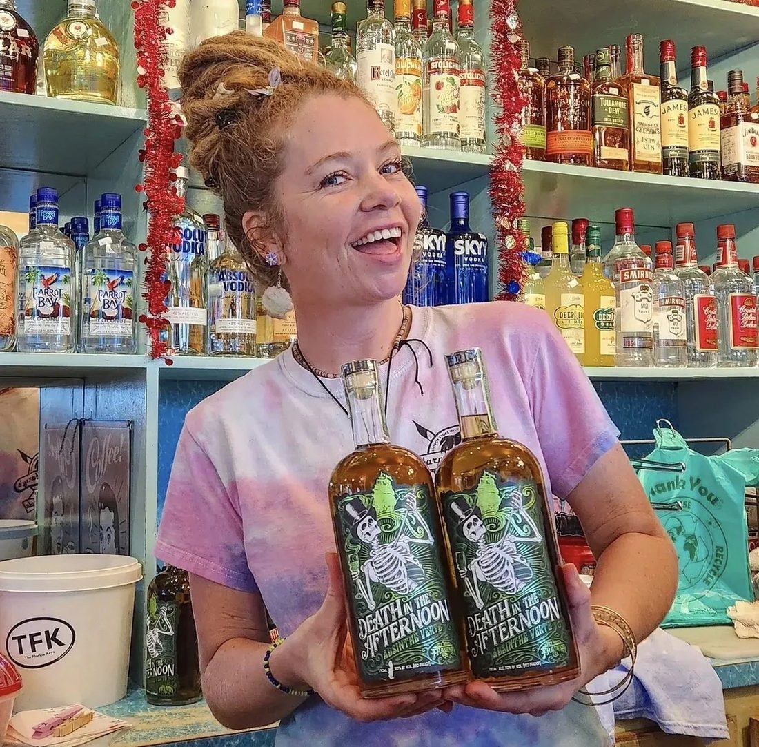 A smiling woman behind a bar holding two bottles of Death in the Afternoon Absinthe.