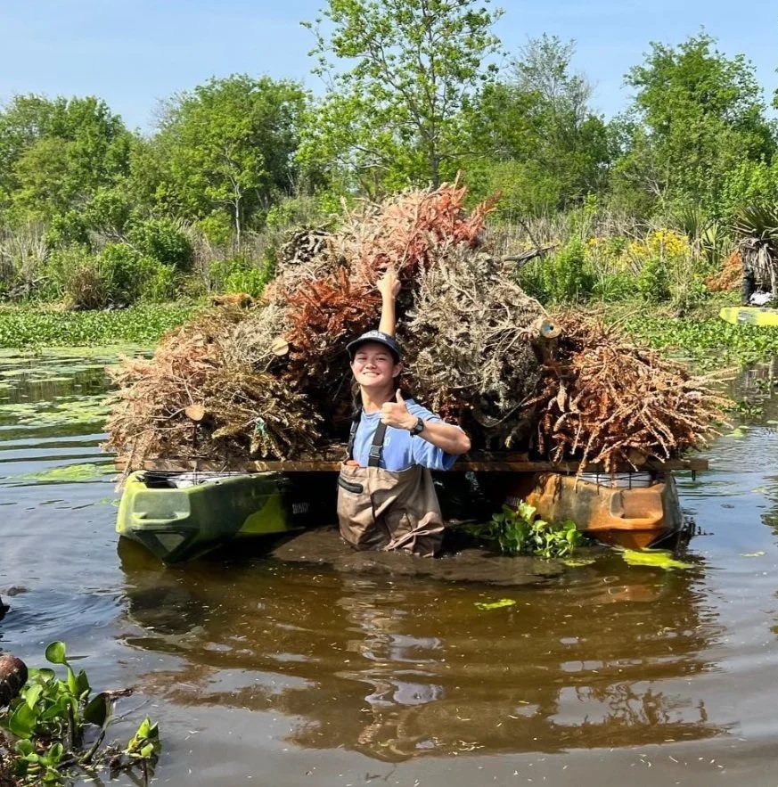 A small boat loaded high with marsh grass on calm water, with a person aboard and trees behind.