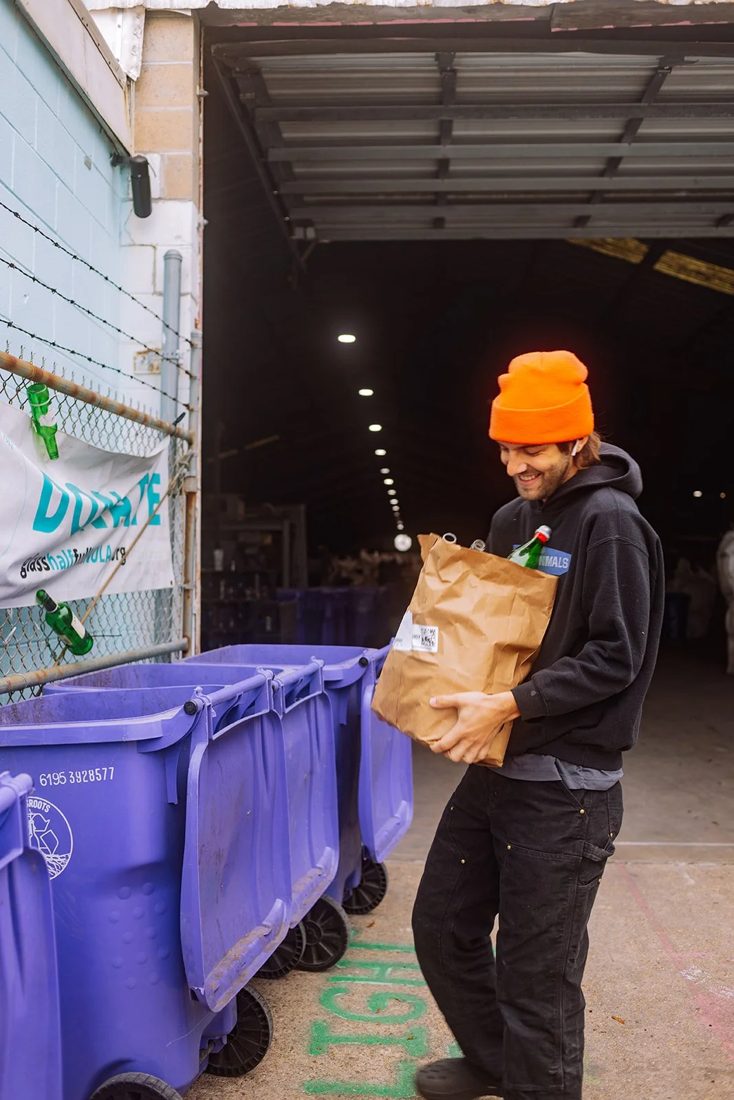 A person in an orange beanie carrying a paper bag of glass toward a row of purple Glassroots recycling bins.
