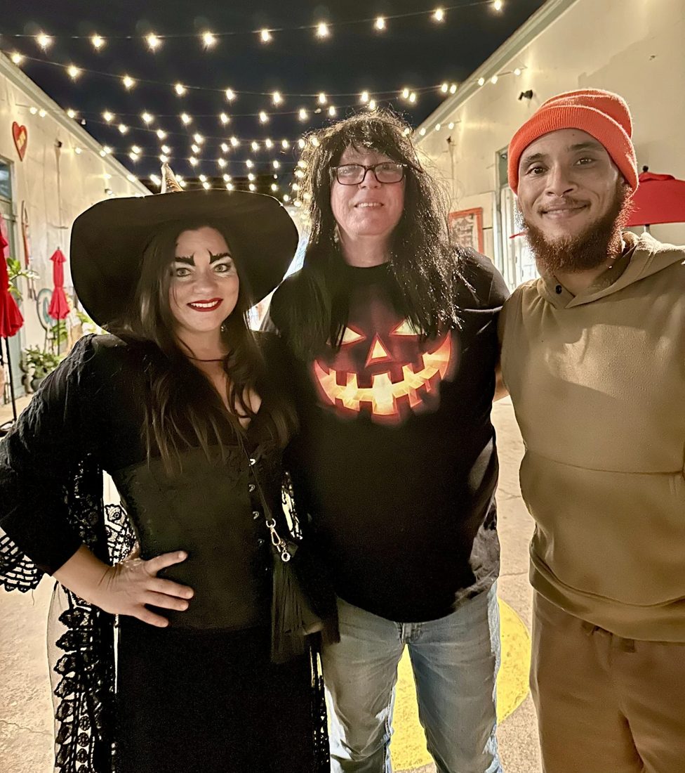 Three people under string lights, one in a witch hat and costume and one in a glowing jack-o'-lantern shirt.